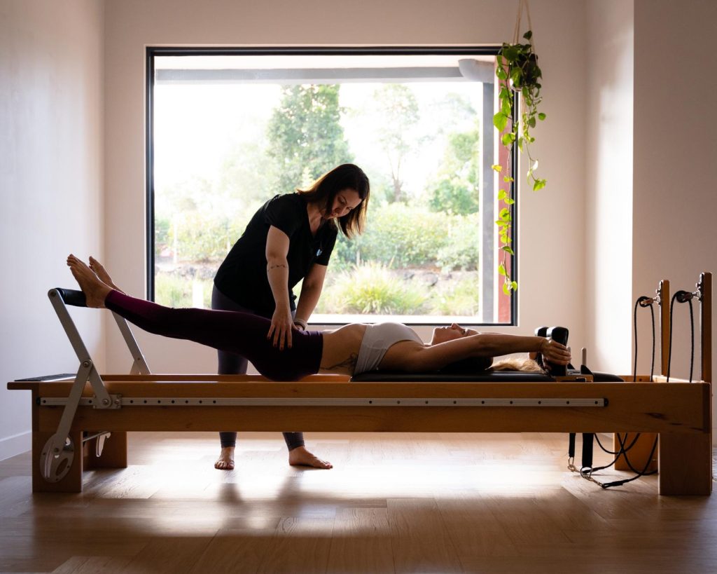 Physiotherapist assisting client on reformer Pilates machine in bright studio, emphasizing rehabilitation and fitness benefits of Pilates classes.